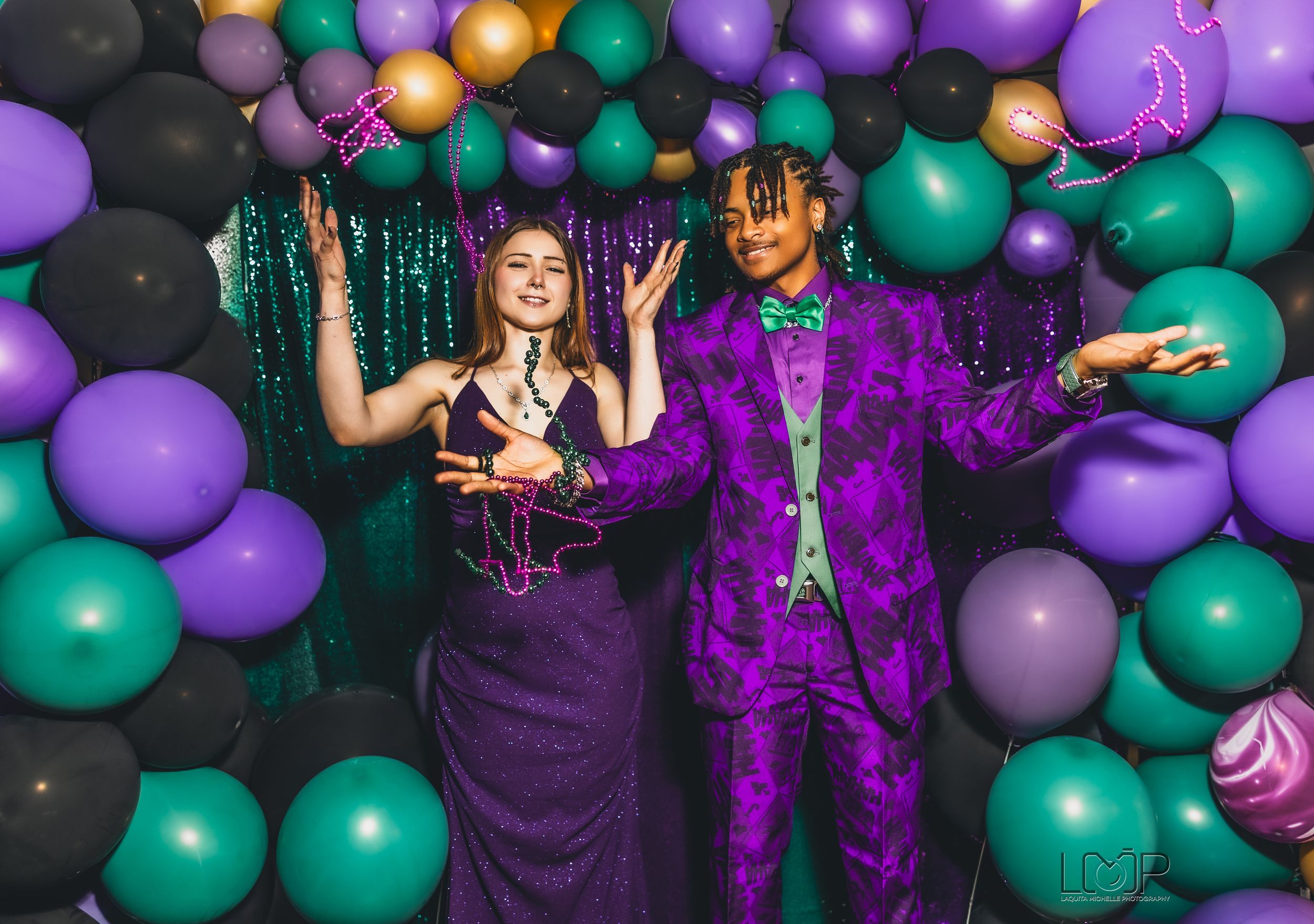 Two people pose at a party against a backdrop of colorful green and purple balloons with sparkly decorations.