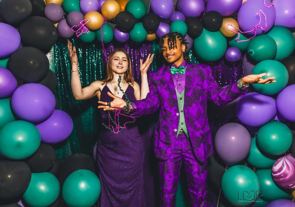 Two people pose at a party against a backdrop of colorful green and purple balloons with sparkly decorations.