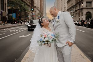 A wedding couple poses in an urban setting with bouquet and formal attire on a city street.