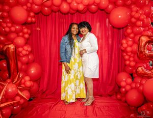 A romantic celebration setup with vibrant red balloon arches and decorative backdrops surrounds a couple in matching outfits.