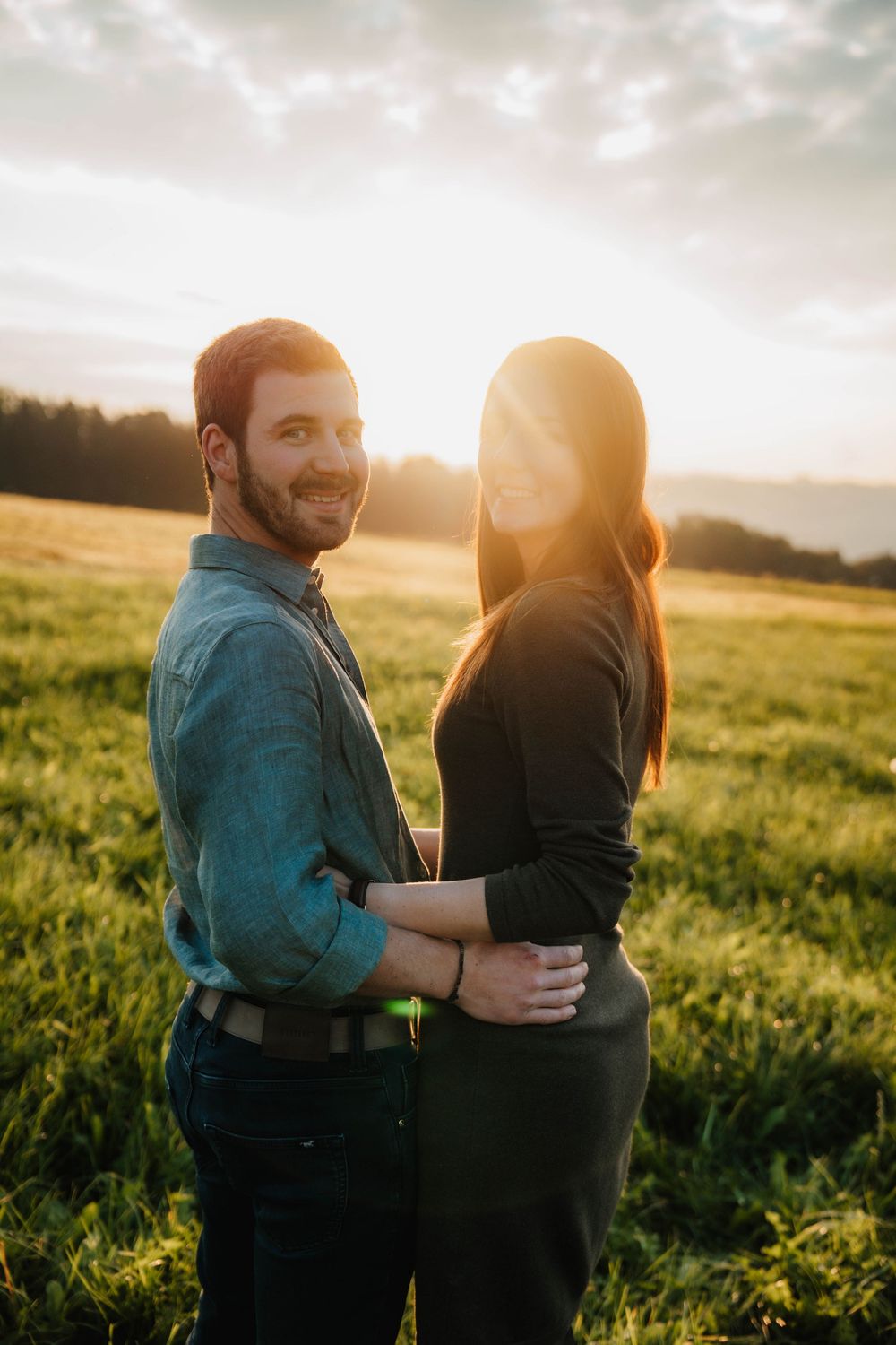 A couple embraces in a sunlit field at golden hour with dramatic cloudy skies overhead.