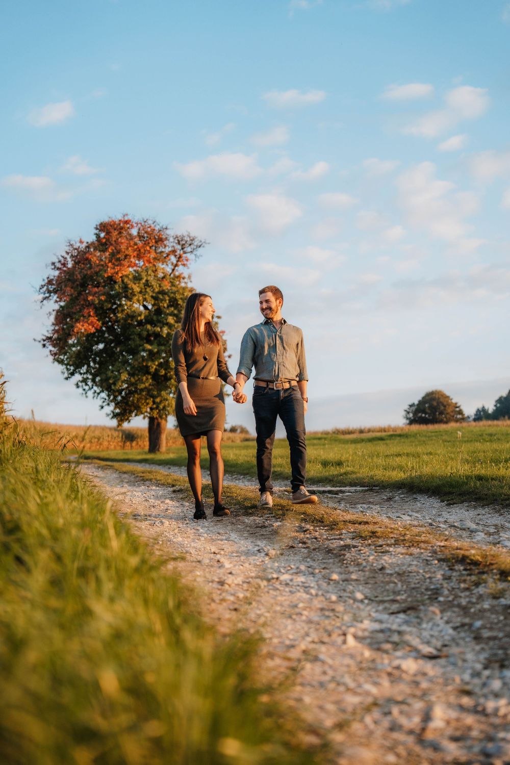 Two people walk together on a dirt path surrounded by autumn foliage and blue skies.