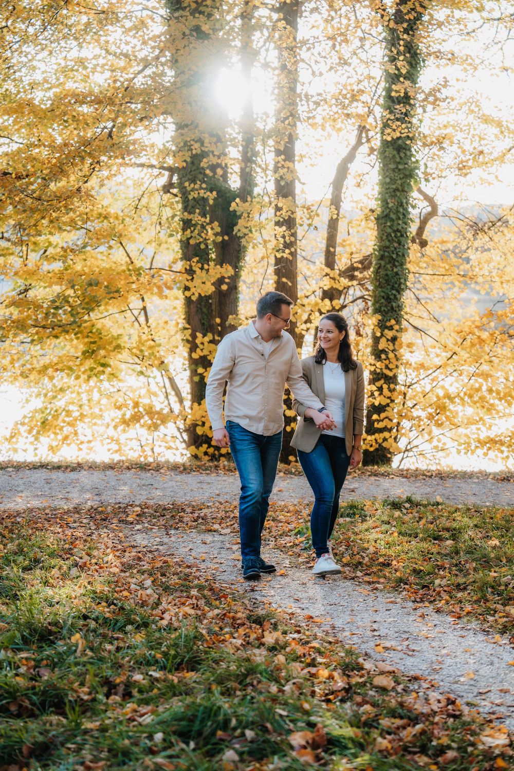 A couple walks together on a leaf-covered path through an autumn forest with golden foliage.
