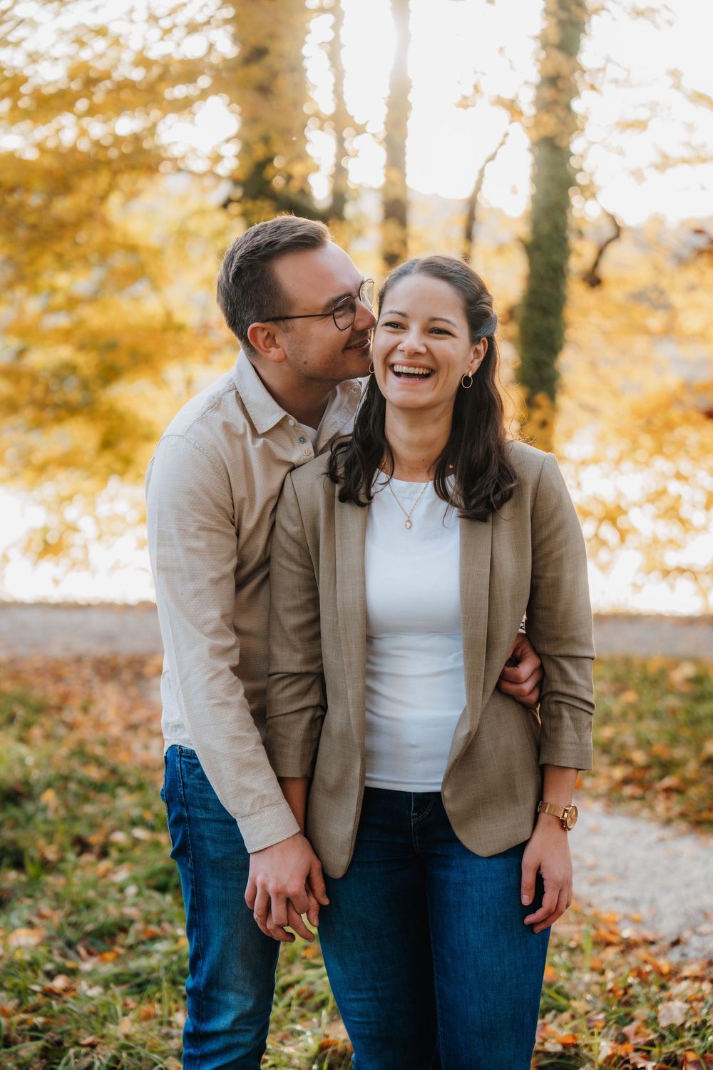 Couple laughing together in autumn foliage wearing coordinated outfits of tan blazer and jeans.