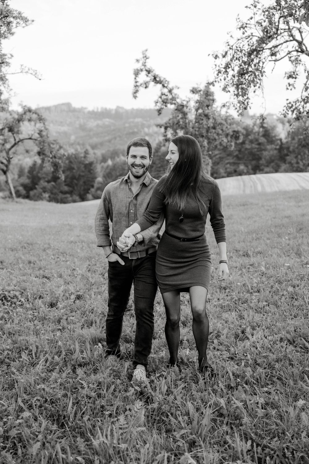 Black and white photo of a couple playfully interacting in a grassy field with mountains in background.