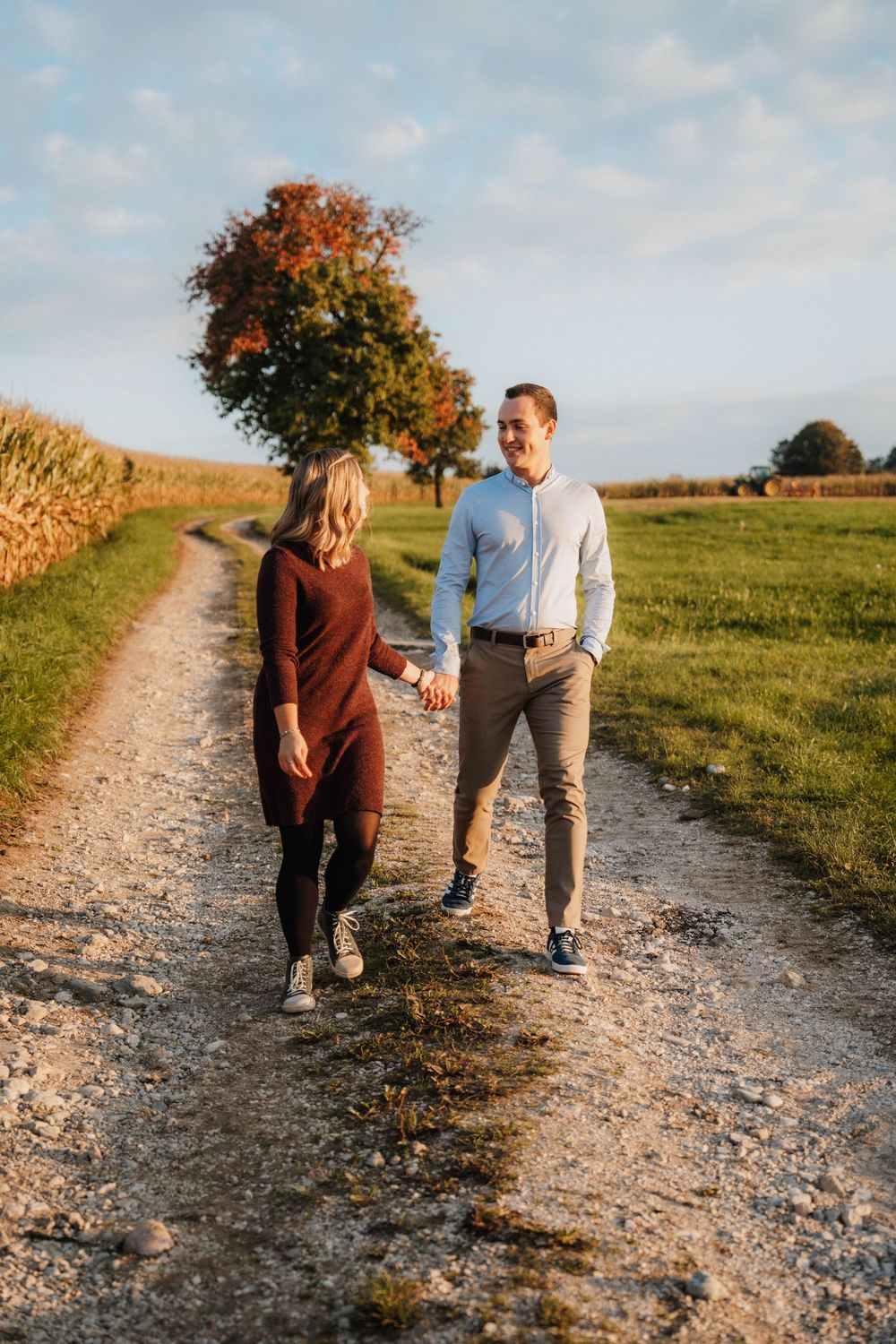 Two people walk together on a gravel path through rolling countryside hills at golden hour.