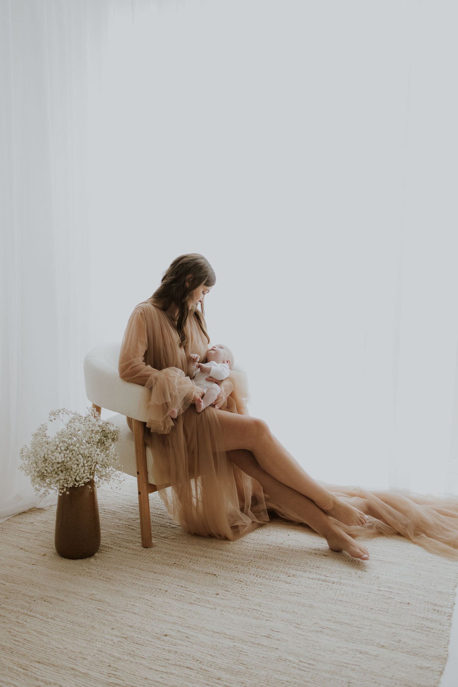 Mother in sheer dress seated with baby in minimalist white room setting.