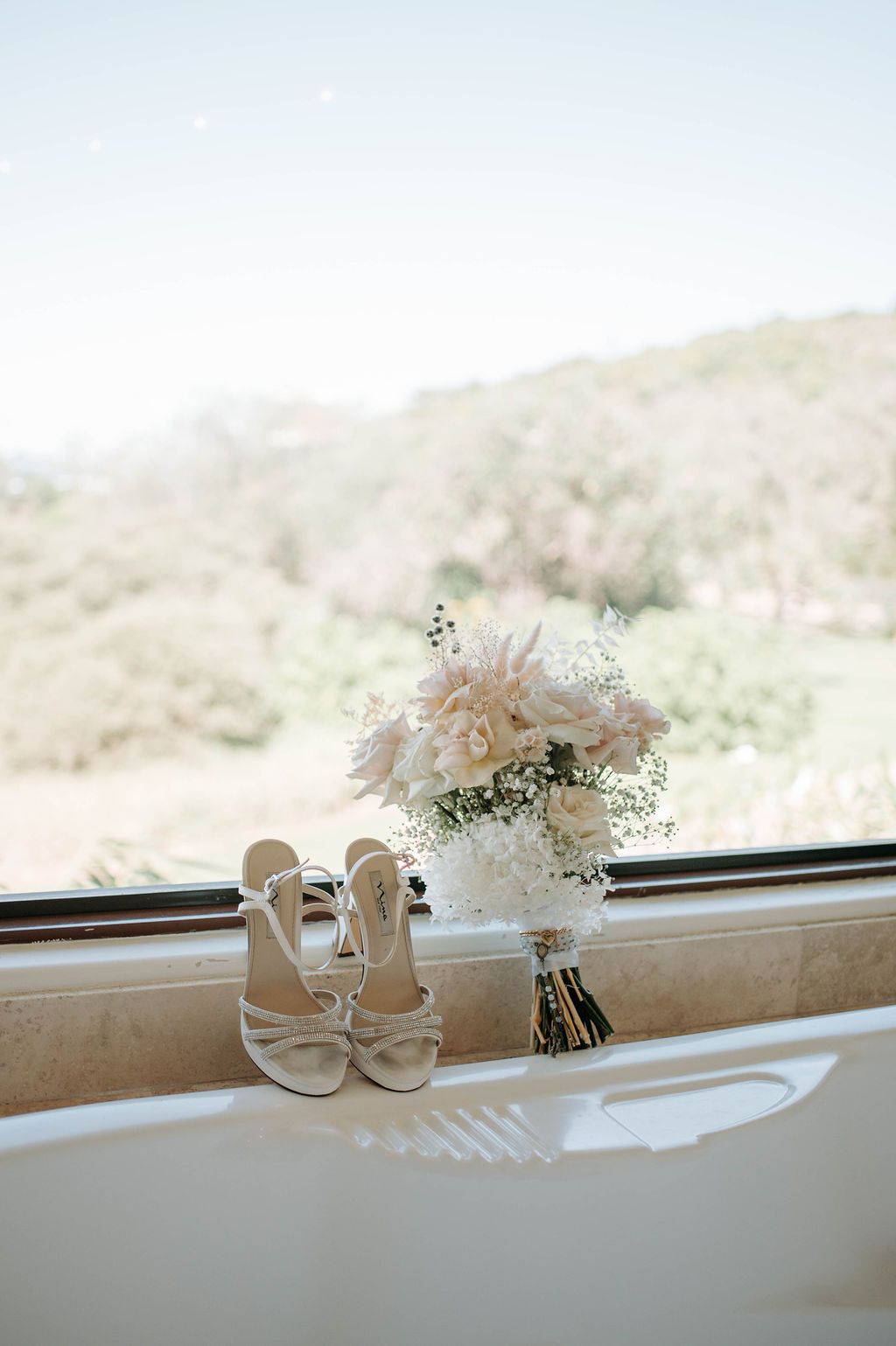 White bridal shoes and bouquet with dried flowers displayed on windowsill with mountain view.