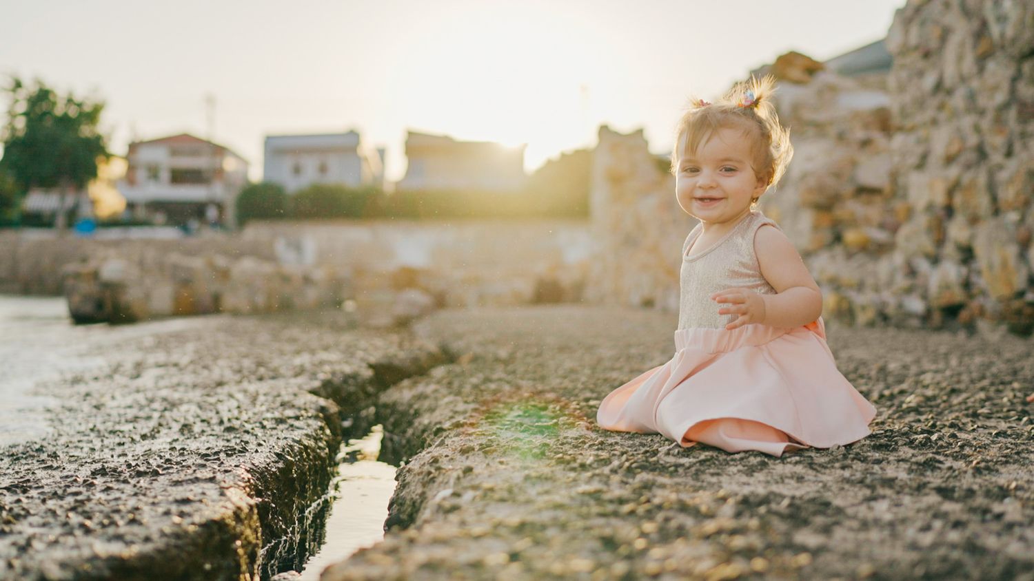 Small child in white dress sitting on rocky ground at sunset.