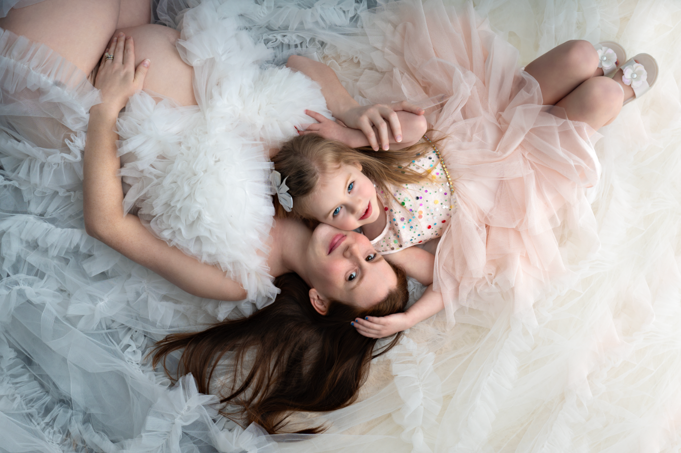 A mother and young daughter in white and pink tulle dresses lying together, sharing a tender moment. Embracing Life’s Beauty