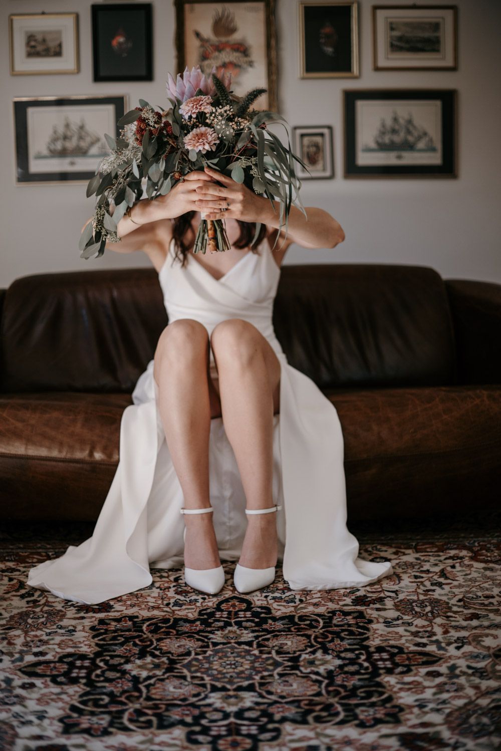 Bride in white wedding dress sitting on leather sofa holding bouquet of flowers covering her face.