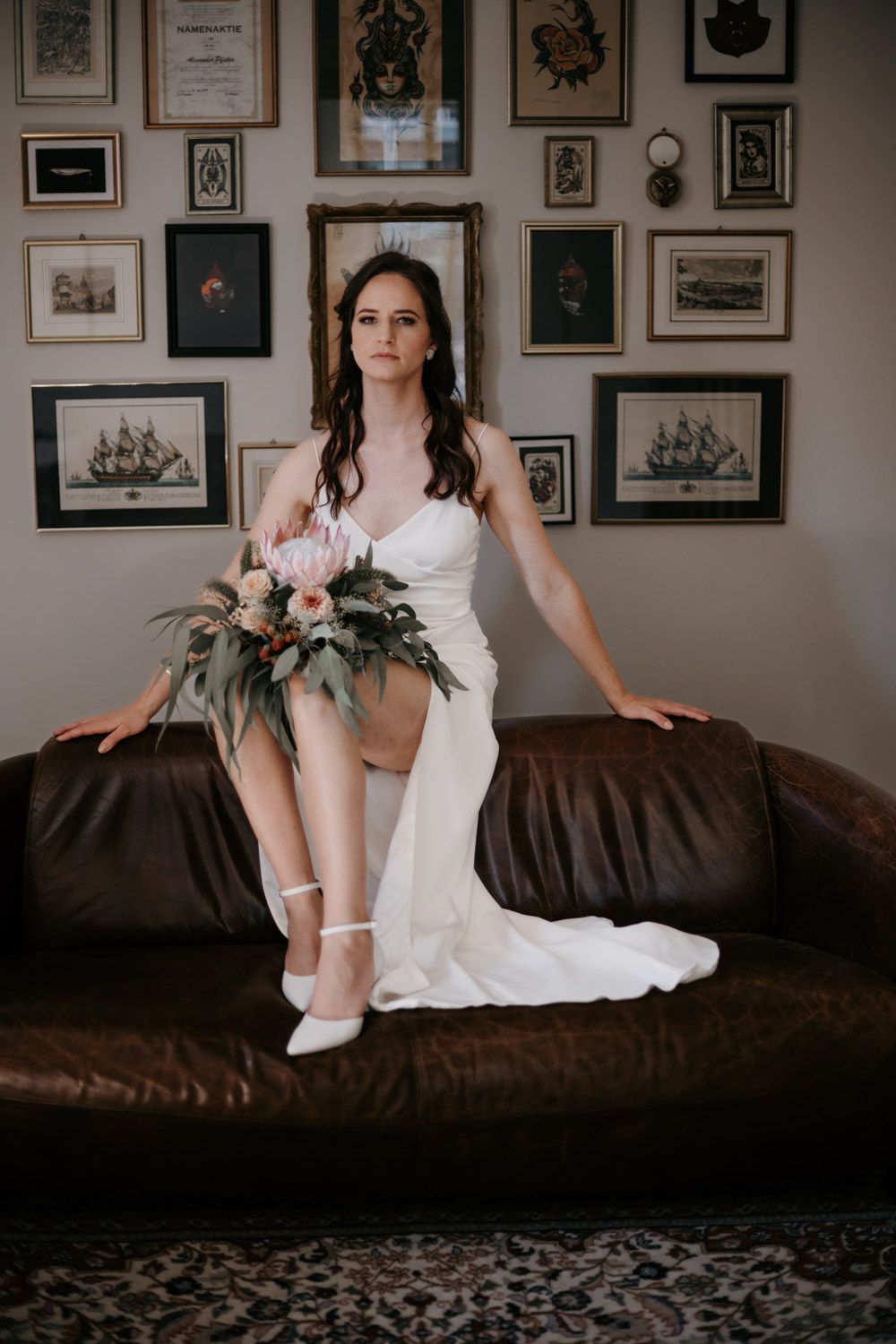 Elegant bride in white gown sitting on leather couch holding bouquet before gallery wall of framed art.