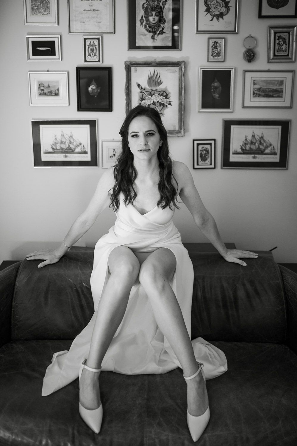 Black and white portrait of bride in white dress seated on leather sofa before gallery wall.