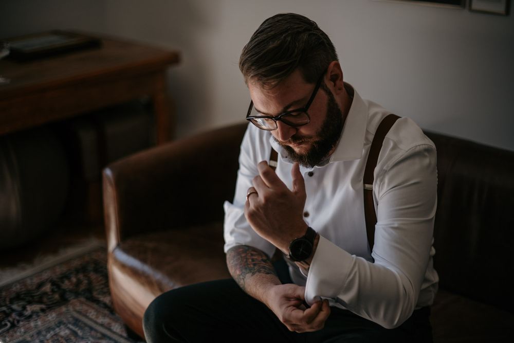 Groom in white shirt and suspenders adjusting cufflinks while seated on a leather sofa.