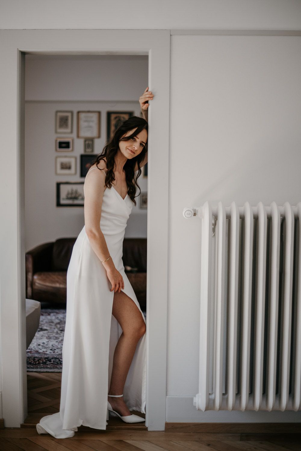 Bride in sleek white wedding dress leaning against doorframe smiling in a bright modern interior.