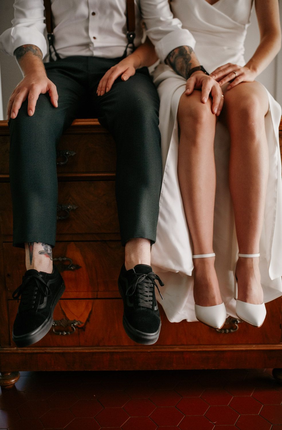 Close-up of bride and groom sitting on dresser, groom in black Vans, bride in white heels.
