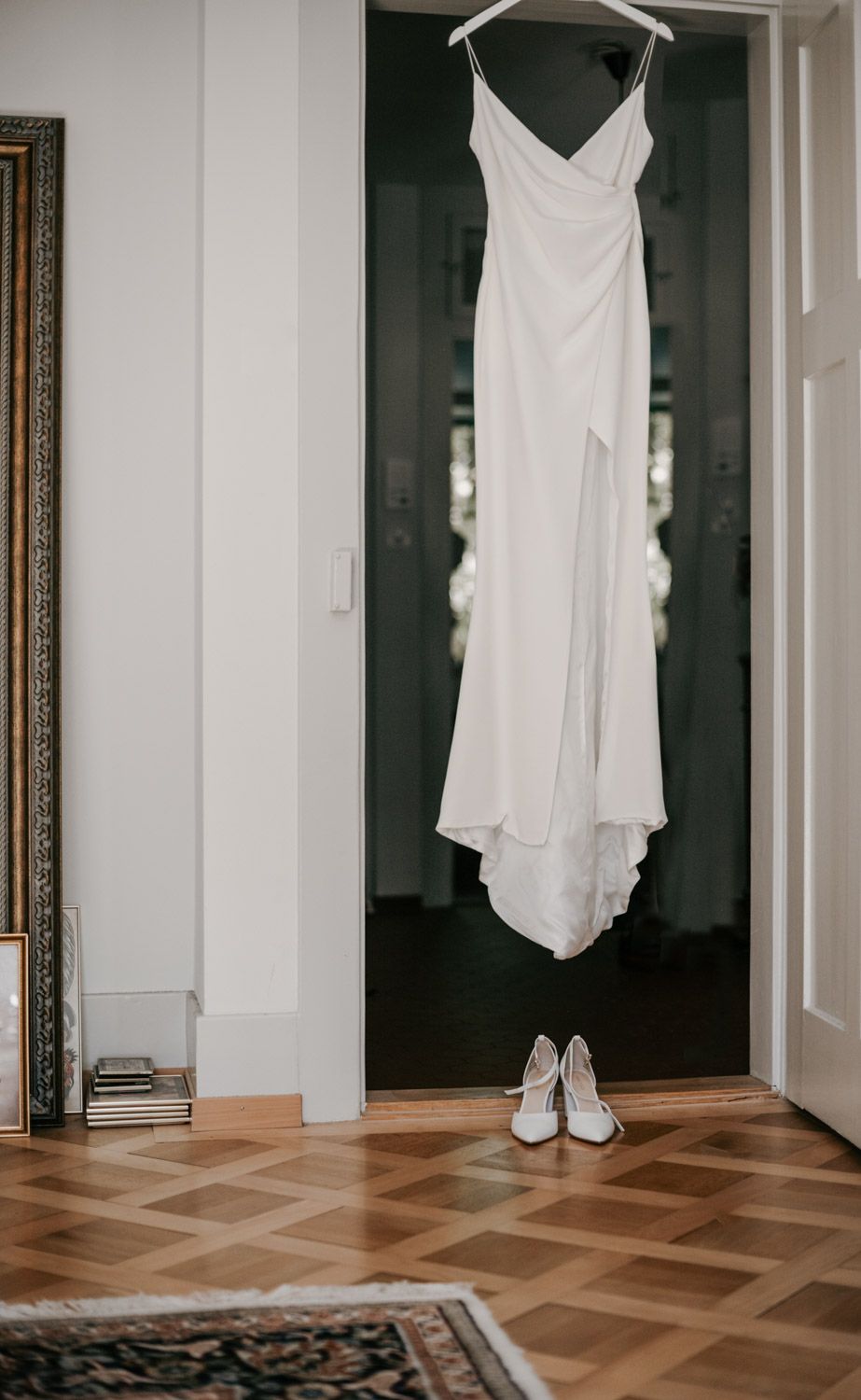 Simple white wedding dress hanging on a door with white heels below on herringbone wood floor.