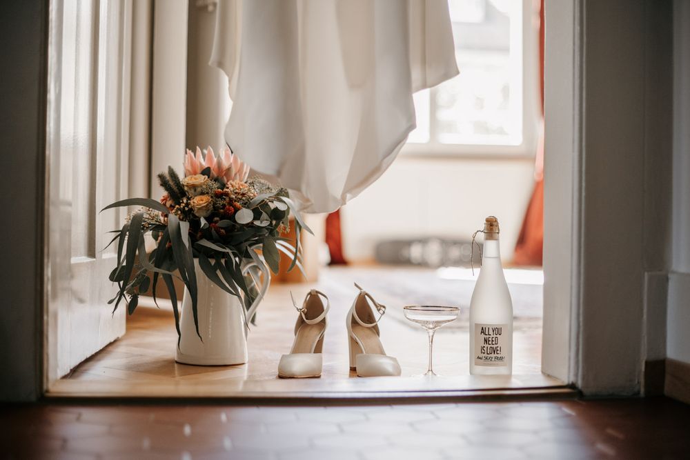 Wedding flat lay with bridal shoes, floral bouquet, champagne glass and bottle arranged by a sunny window.