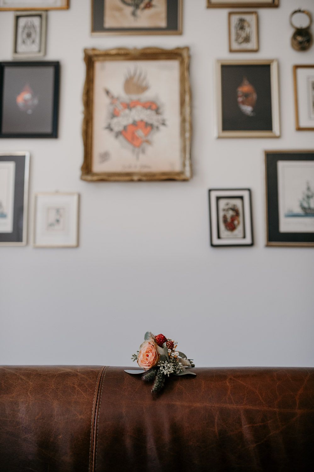 Small floral boutonniere resting on brown leather sofa beneath eclectic gallery wall of framed tattoo artwork.