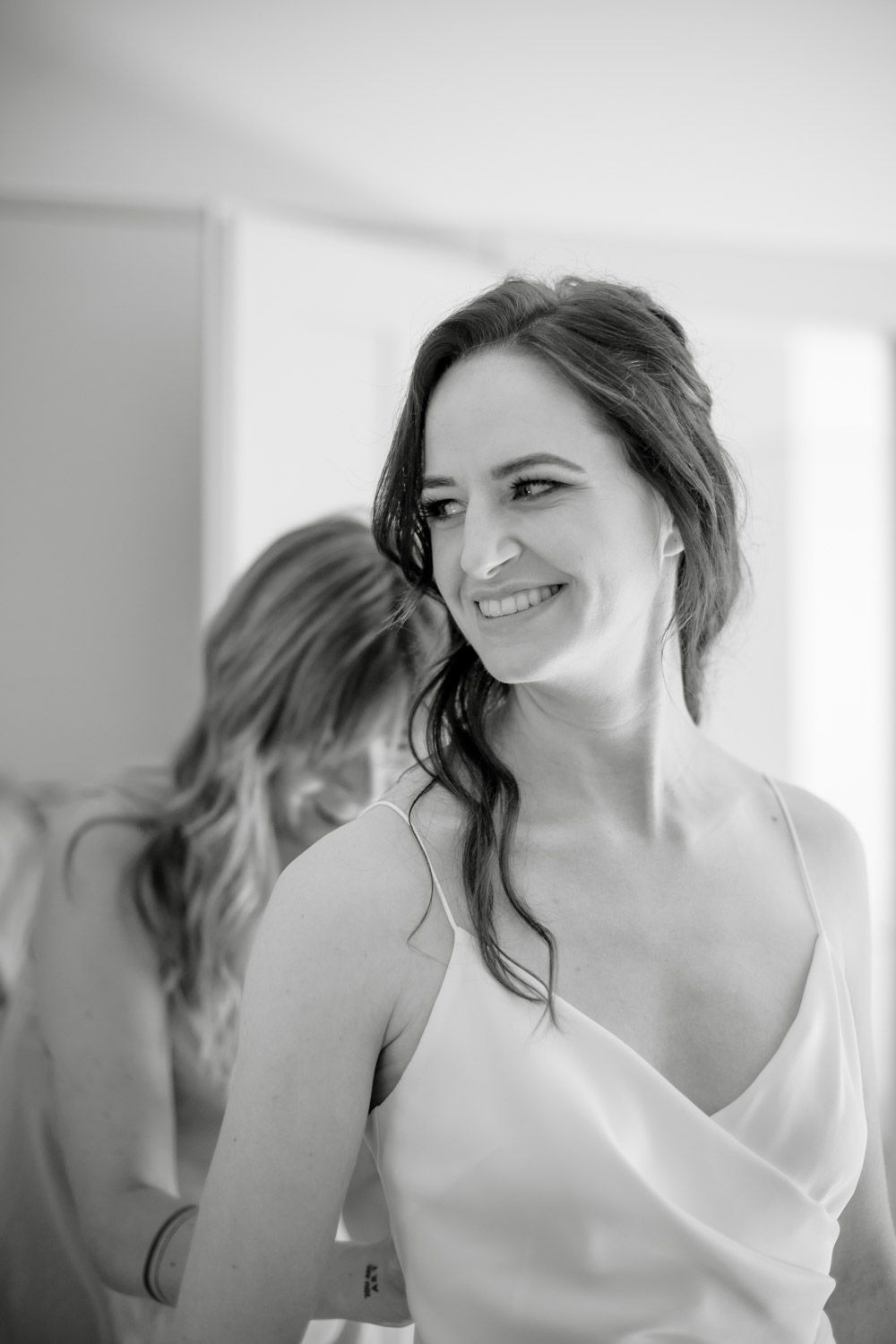 Smiling bride in white spaghetti strap gown getting ready, black and white portrait with soft natural light.