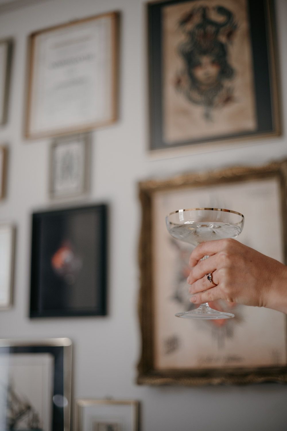 Hand holding elegant coupe glass with gold rim in front of eclectic gallery wall of framed artwork.