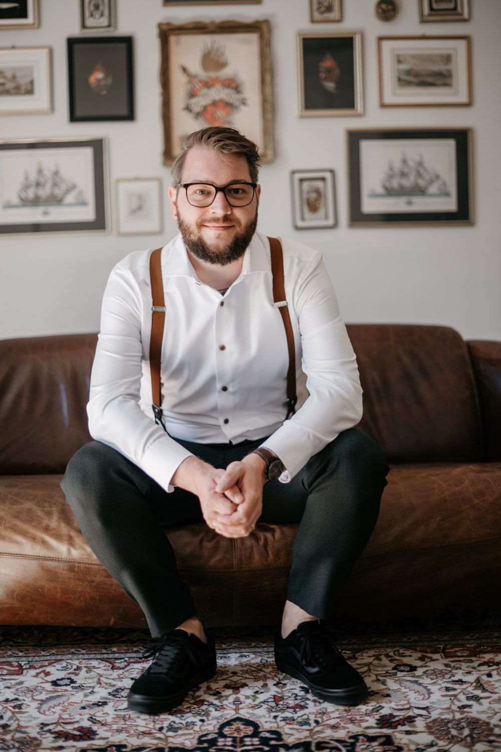 Bearded groom in white shirt and brown suspenders sitting on leather sofa beneath gallery wall of framed prints.
