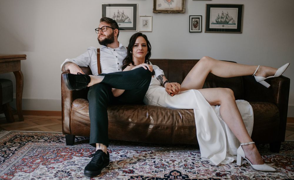 Bride and groom sitting casually on a brown leather sofa in a stylish room with framed artwork on the wall.