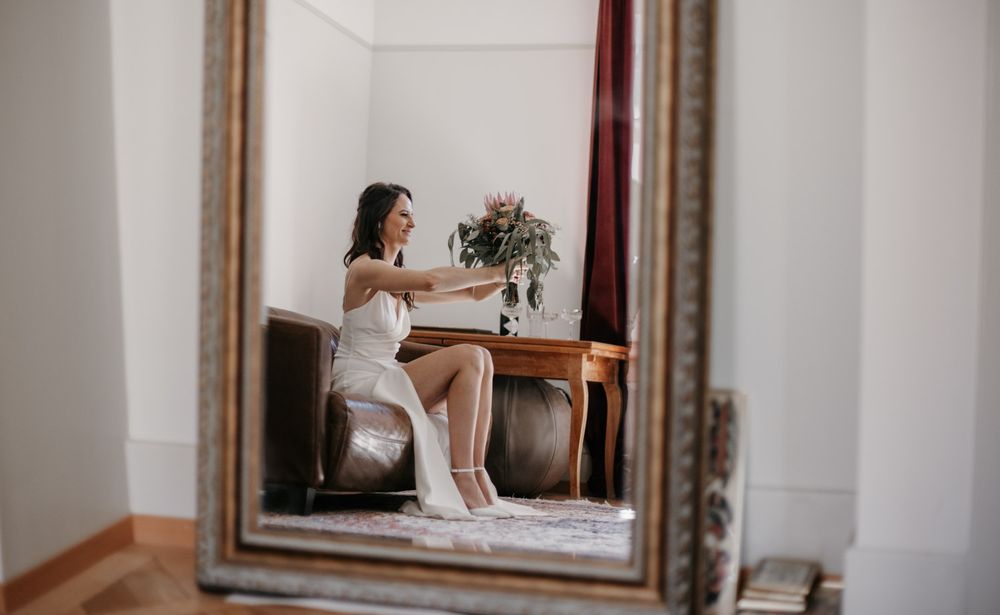 Bride in white dress sitting before a mirror, holding a bouquet in a softly lit elegant room.