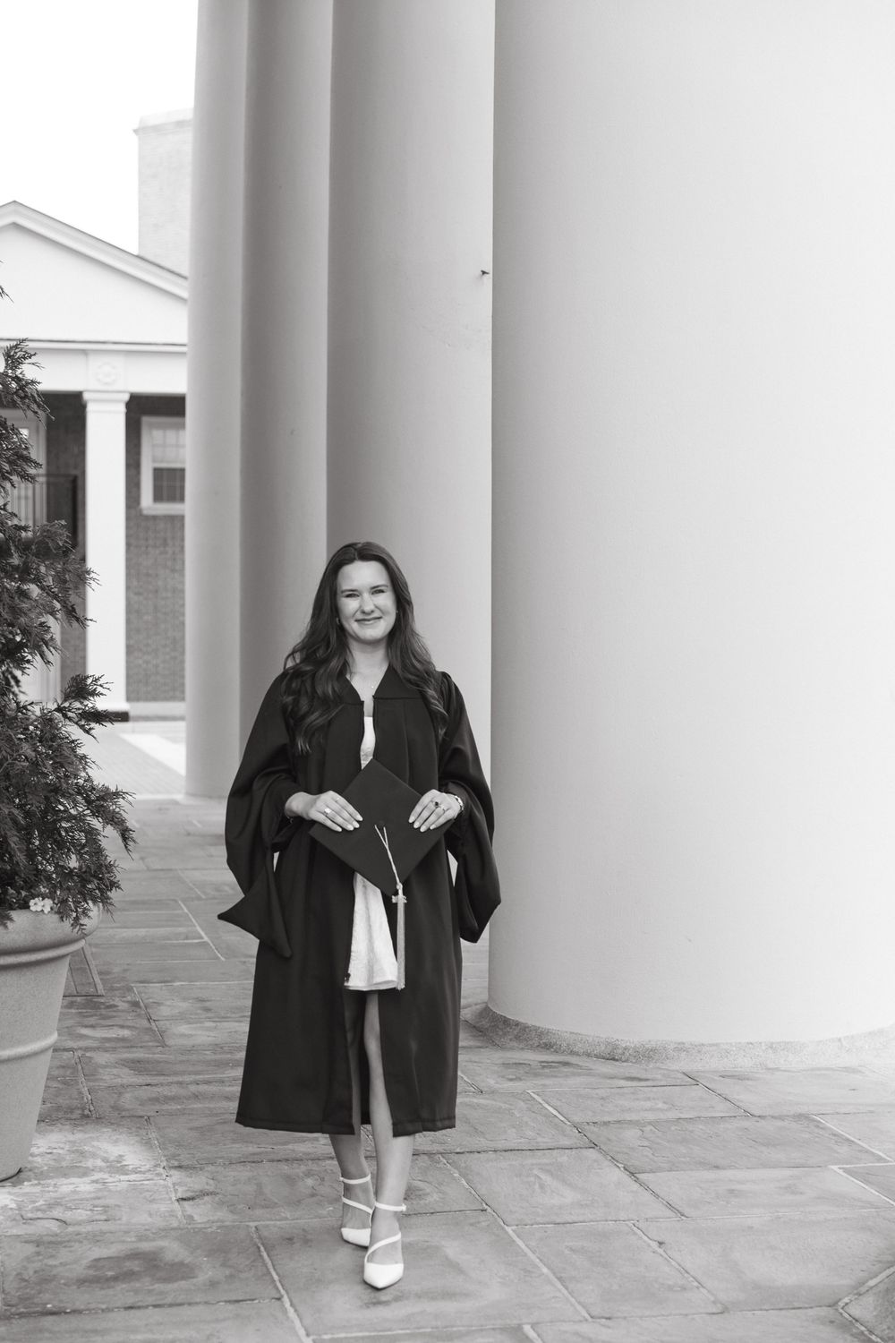 Black and white photo of graduate in academic regalia standing by white column.