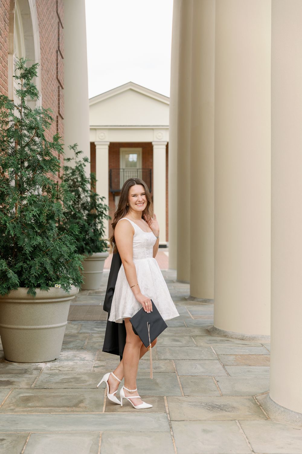 Graduate in white dress and graduation gown poses by stone columns and potted plants.