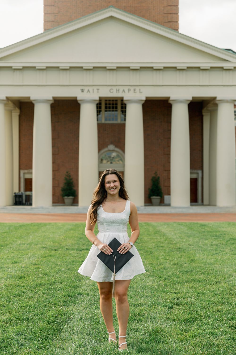Graduate in white dress poses in front of Wait Chapel with diploma on sunny day.