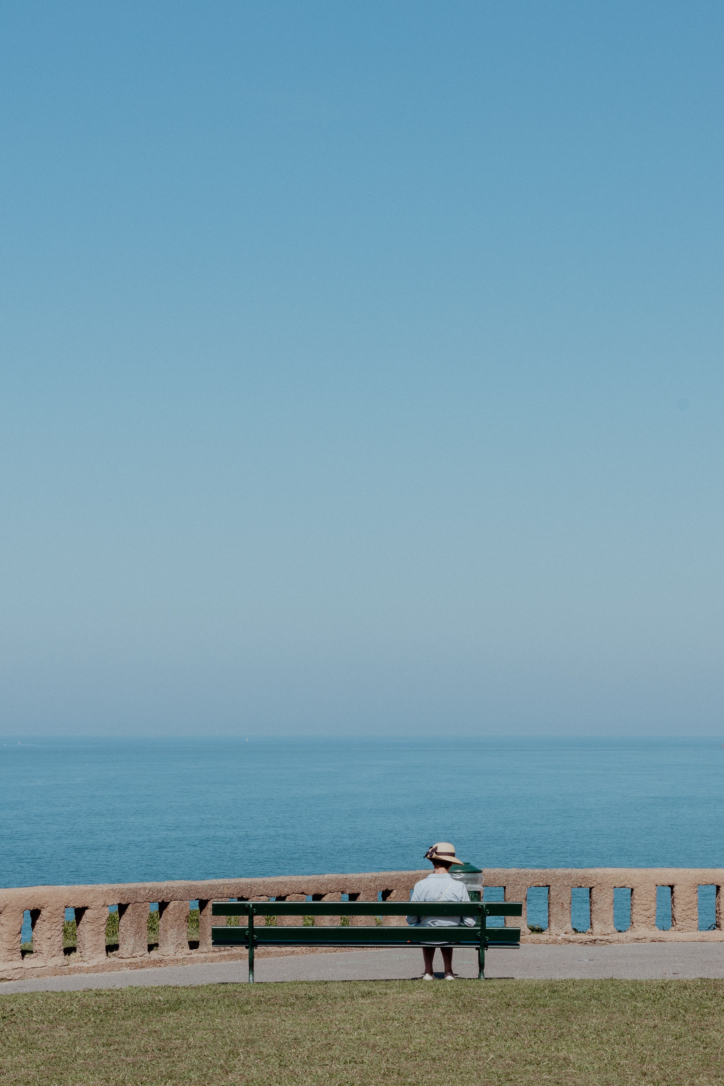 Person sitting alone on bench overlooking vast ocean view under clear blue sky.