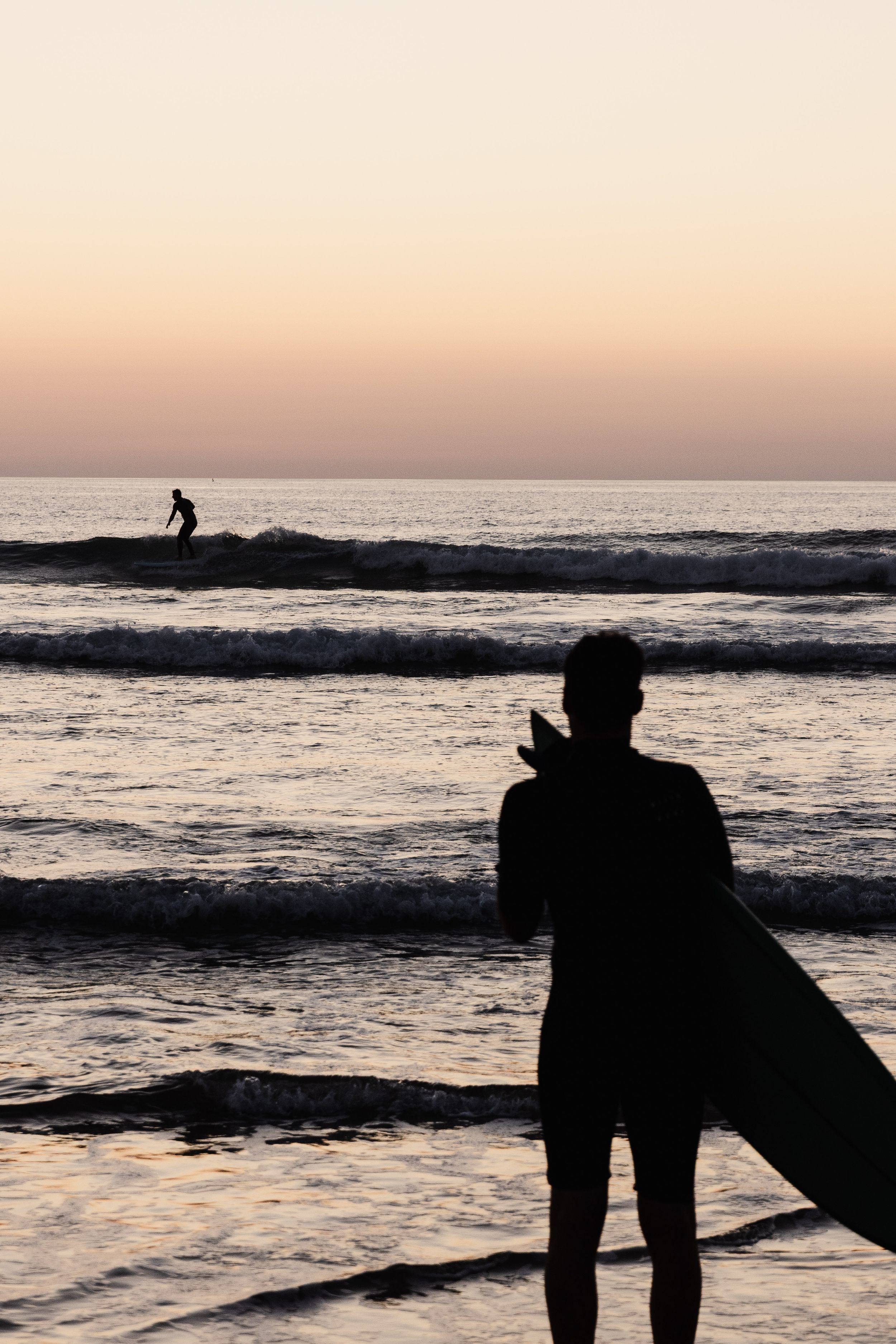 Silhouette of a surfer standing on the beach at sunset while watching waves roll in.