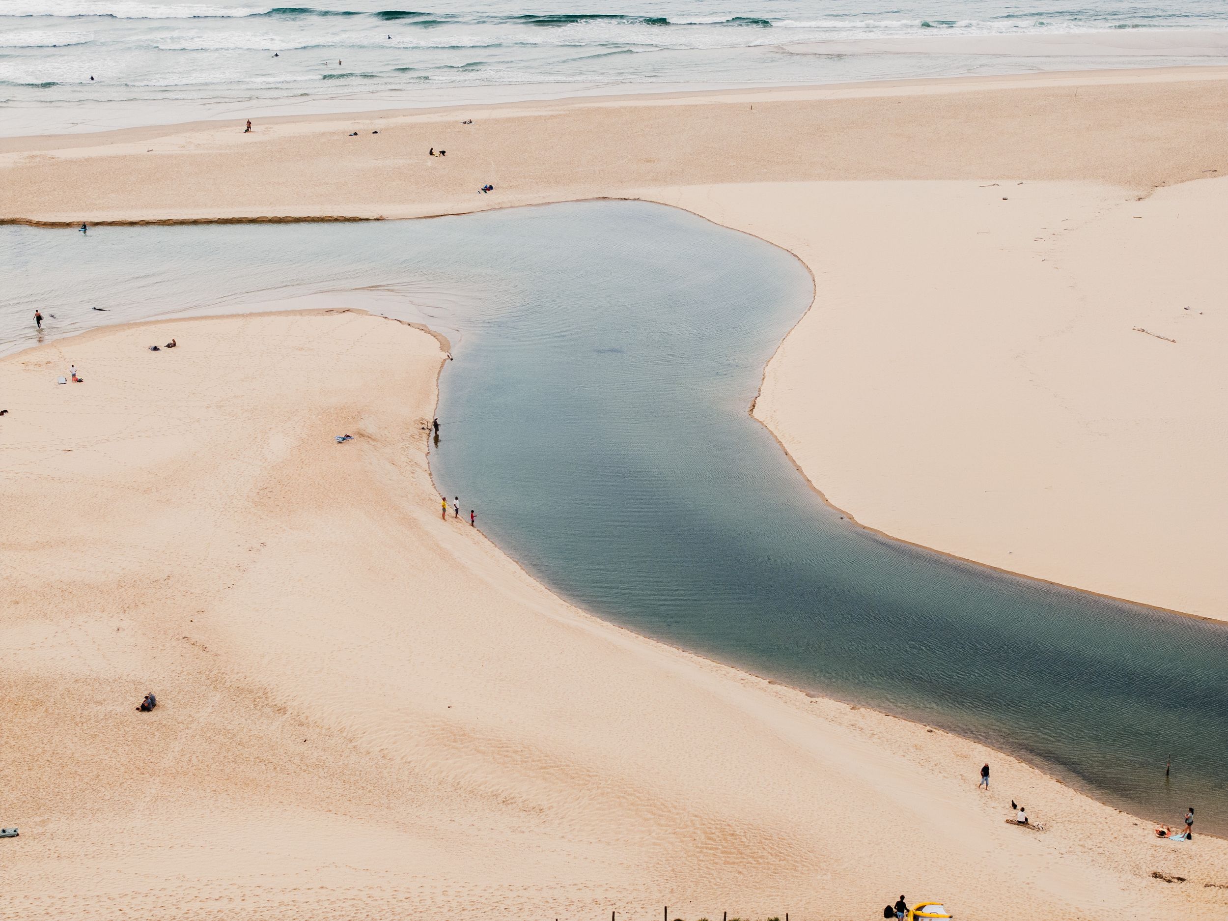 Curved river inlet meeting sandy beach viewed from above with tiny beachgoers.