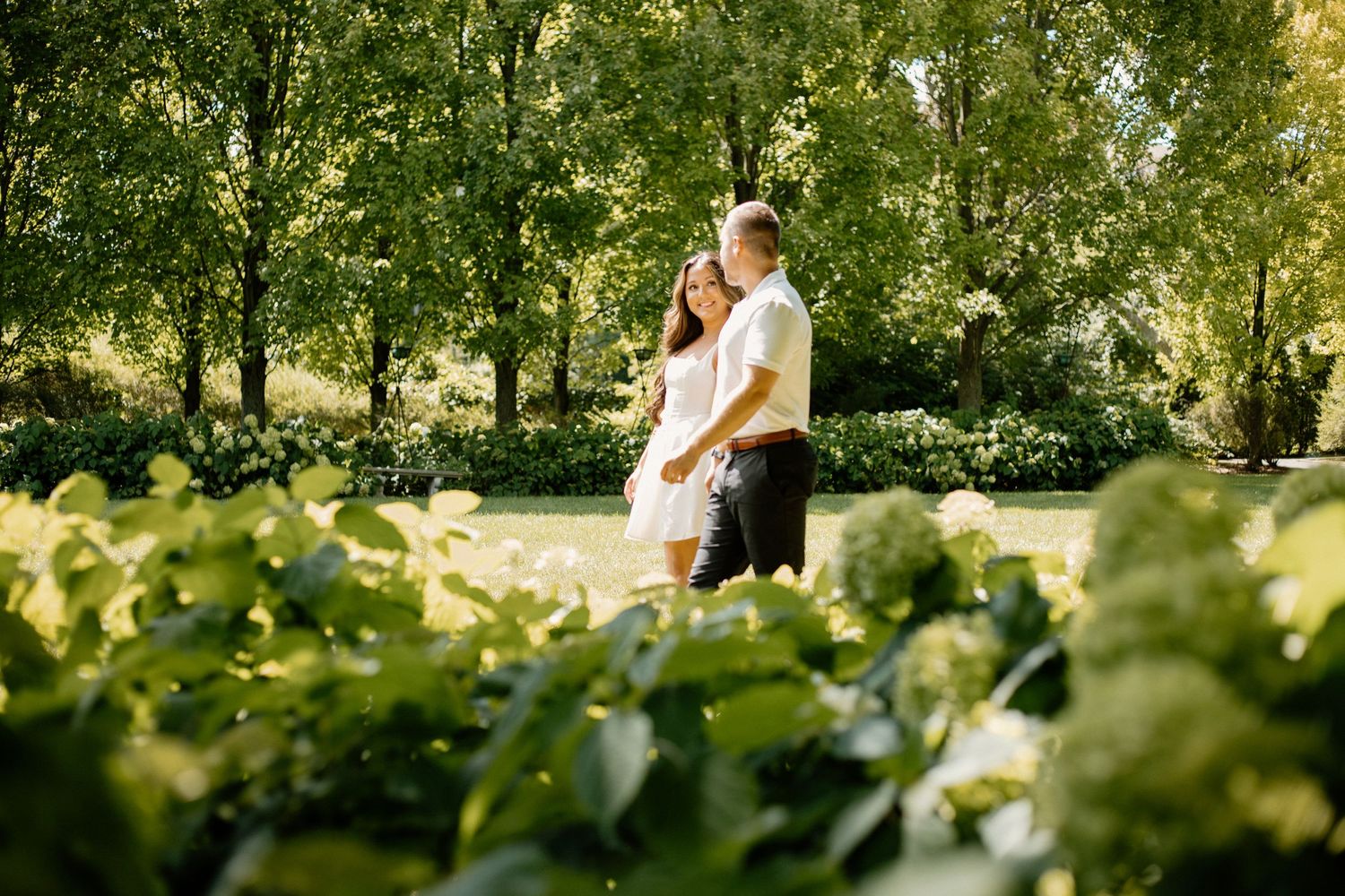 A romantic couple embraces in a lush green garden setting with sunlit trees and foliage in the background.