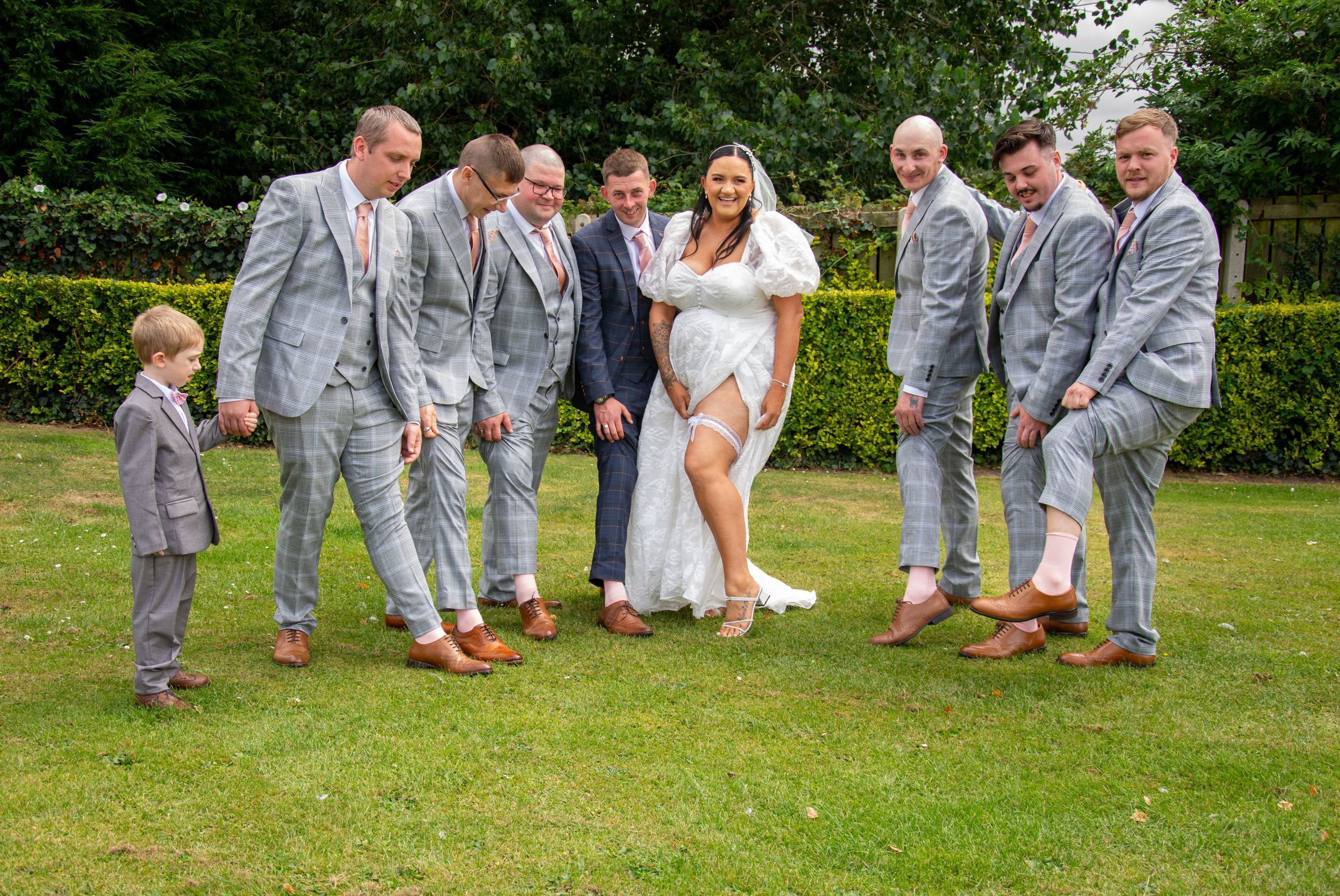 Fun wedding party photo with groomsmen in grey suits on manicured lawn.