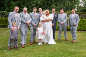 Wedding party in matching grey suits poses on manicured lawn during summer celebration.