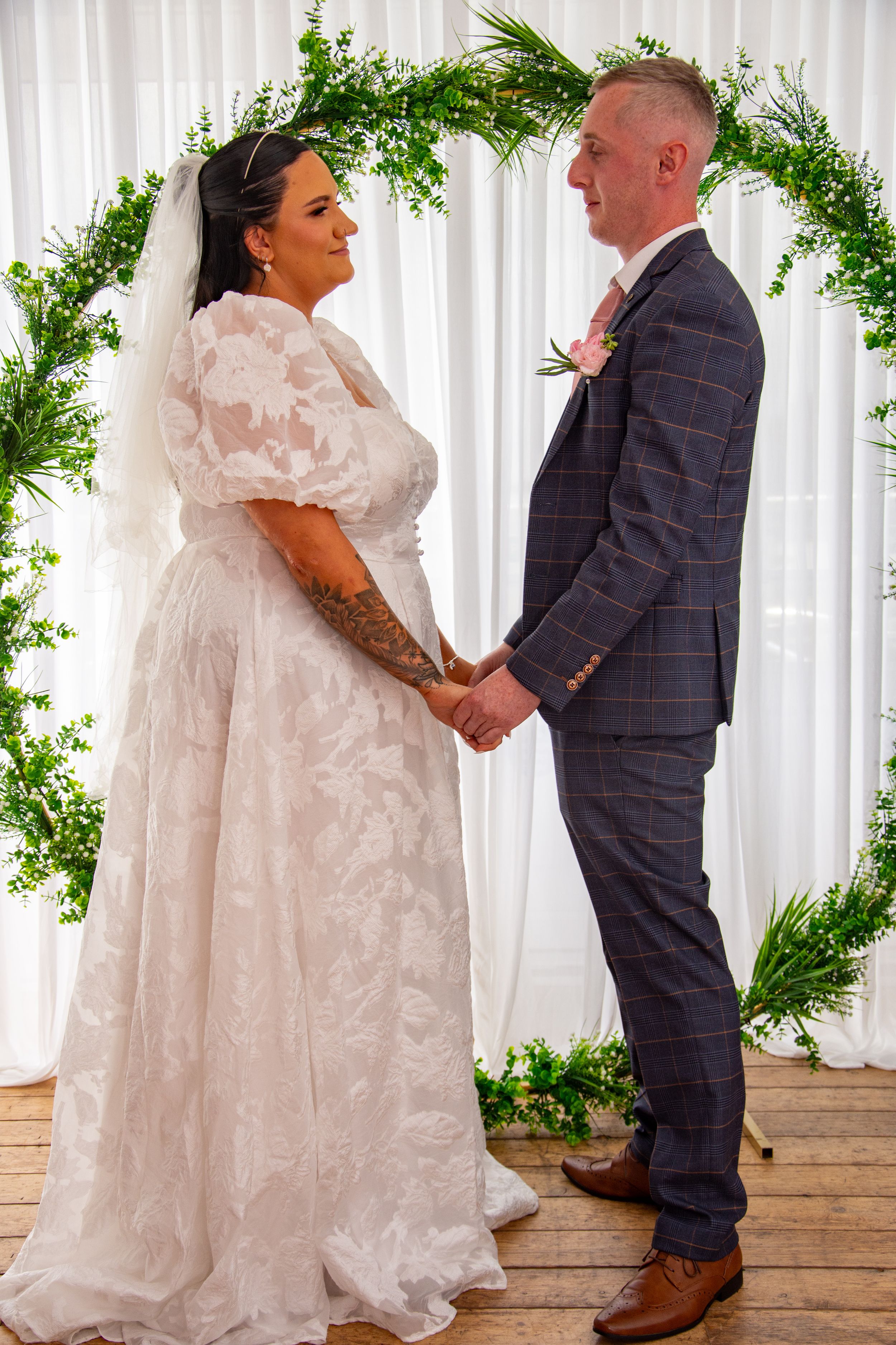 Couple exchanging vows beneath a green floral arch at their wedding ceremony.