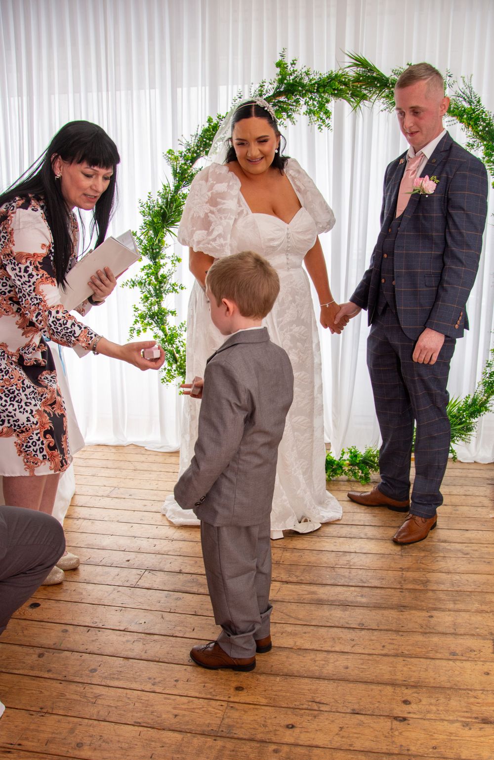 Wedding ceremony taking place under a greenery arch with a young attendee in a grey suit participating.