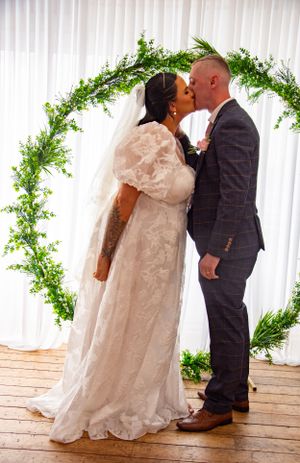 Romantic wedding kiss under an elegant greenery arch against white backdrop.