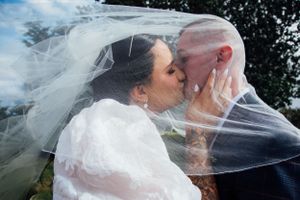 Romantic wedding kiss under flowing white veil against blue sky backdrop.