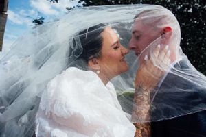 Romantic wedding moment captured through a flowing white veil against outdoor scenery.