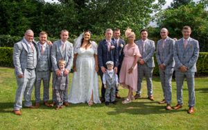 Wedding party in coordinated grey suits poses with couple on lawn.