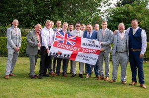 Groomsmen posing outdoors holding a British-themed banner during wedding celebrations.