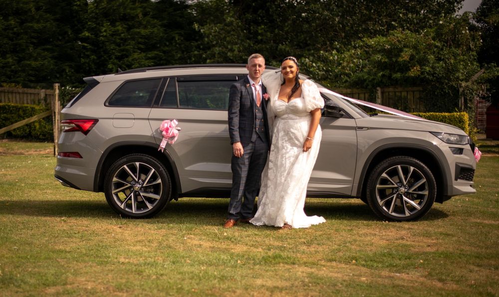 Newlyweds pose beside decorated grey SUV on grass.