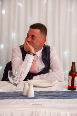 Wedding guest in formal attire sitting at decorated reception table with drinks.