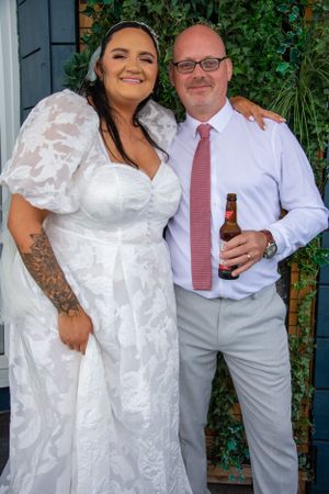Elegant wedding couple poses against ivy-covered wall at outdoor venue.