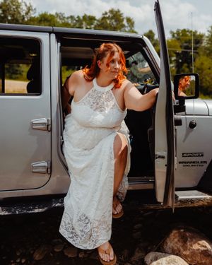 Person in white lace dress exiting silver Jeep in outdoor setting.