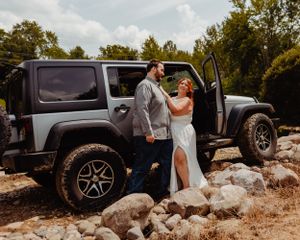 Couple shares intimate moment beside grey Jeep during outdoor wedding photoshoot.