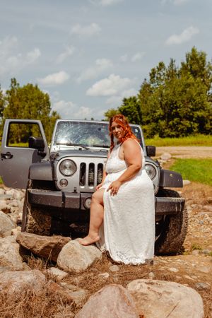 Bride in white lace dress poses on rocks next to muddy Jeep Wrangler.